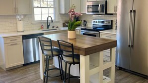 Une cuisine moderne avec des meubles blancs, un îlot central en bois avec deux chaises, des appareils électroménagers en acier inoxydable et un luminaire suspendu.