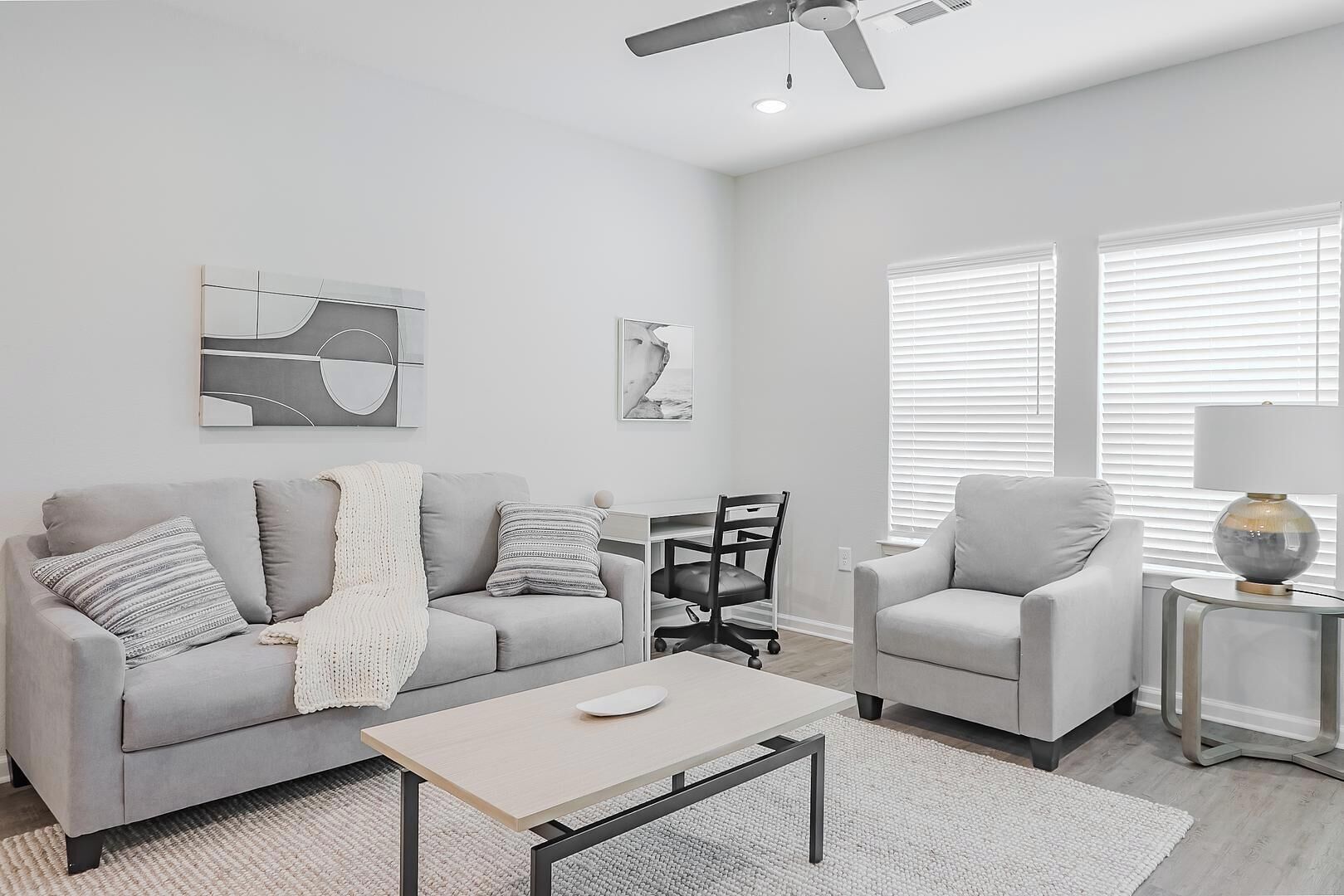 A modern living room with a grey sofa, a small table, and a chair. There are two windows with blinds, a ceiling fan, and a lamp on a side table.