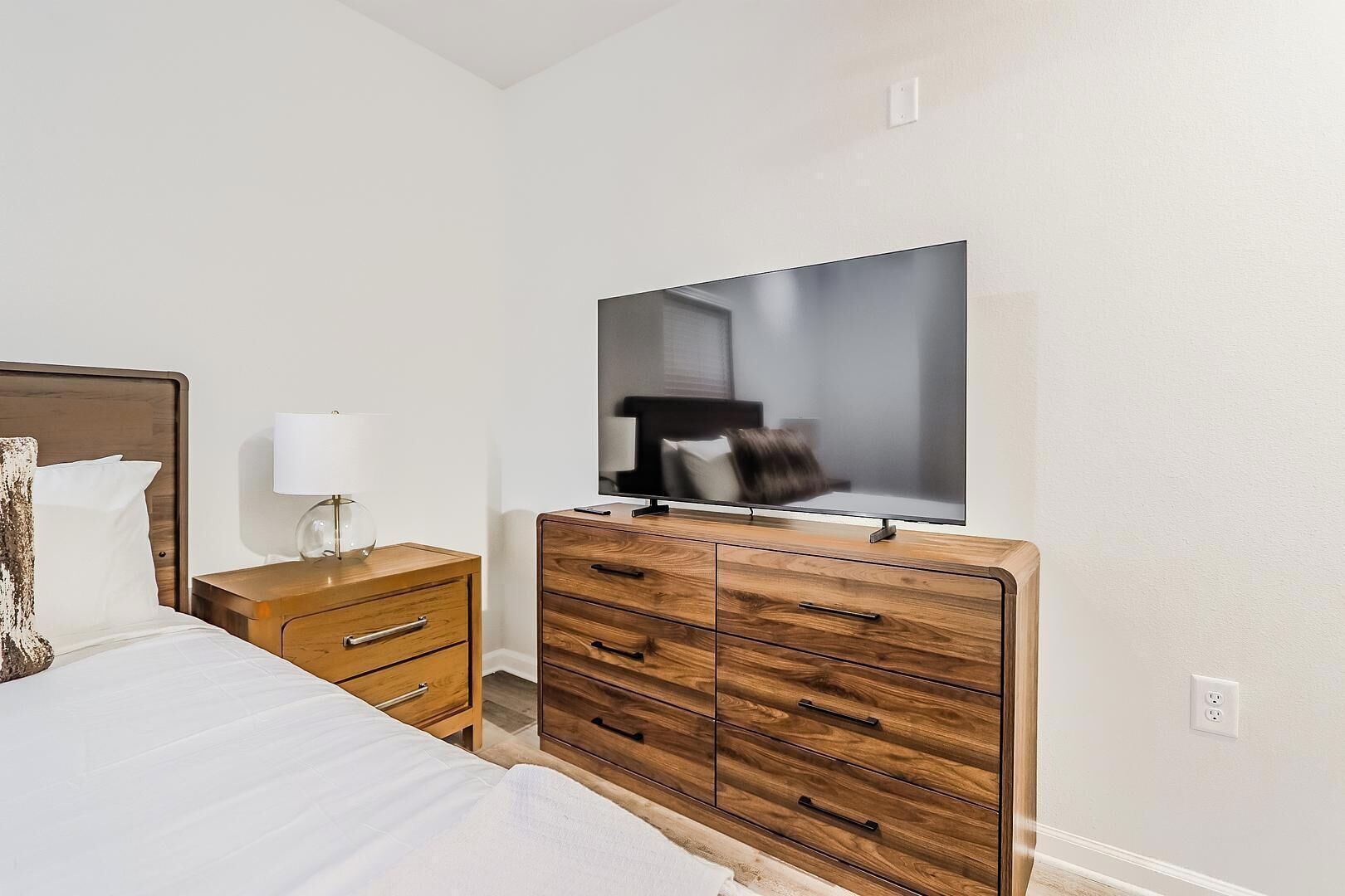 A hotel room with a bed, a wooden dresser with drawers, a TV on the dresser, and a nightstand with a lamp.