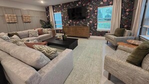 En stue med sofaarrangement, sofabord, sengebord og et fjernsyn ophængt på væggen.