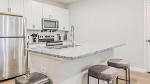 Moderni keittiö, jossa on valkoiset kaapit, ruostumattomasta teräksestä valmistetut kodinkoneet, graniittitaso ja kaksi baarituolia.