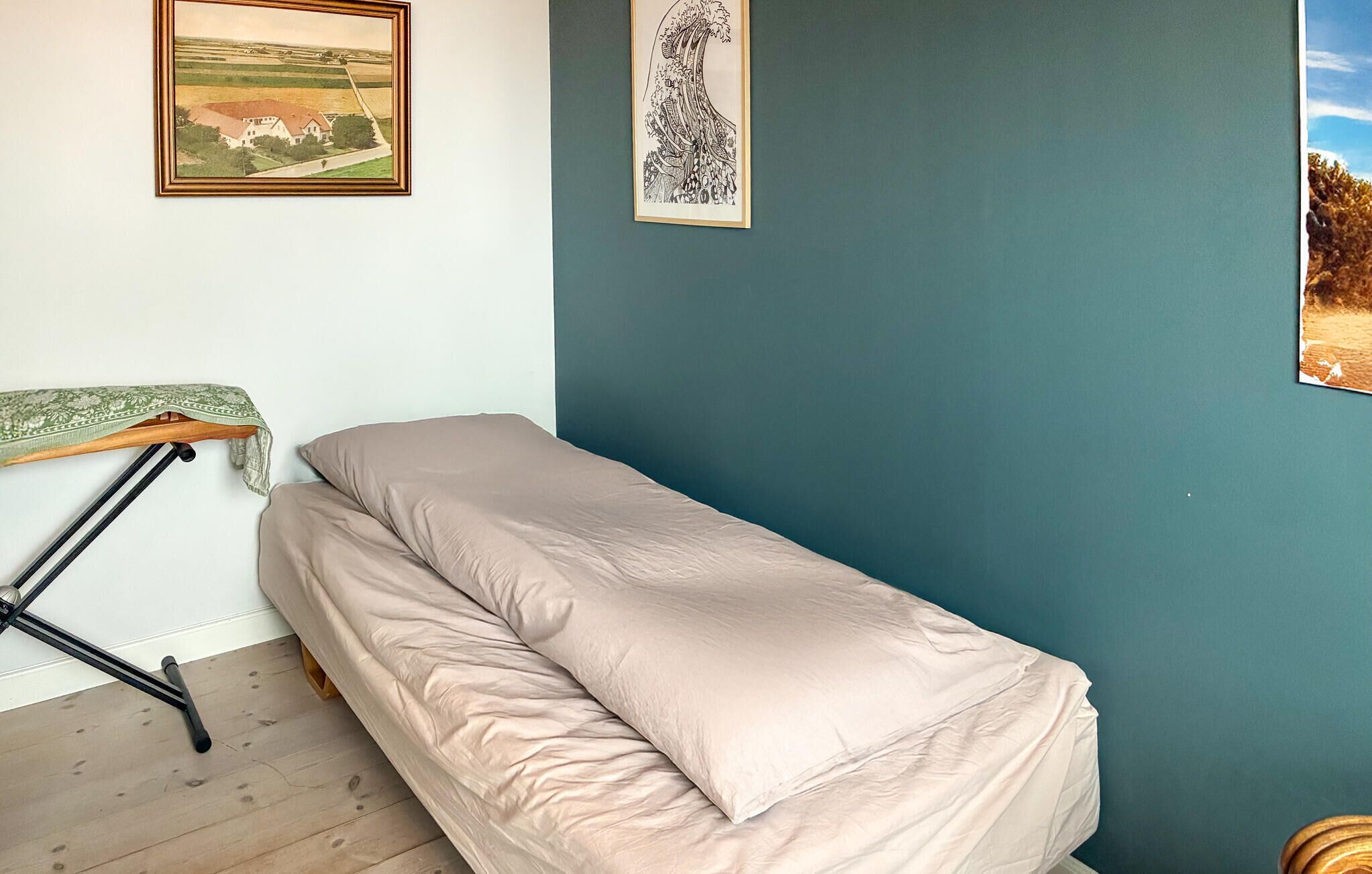 A small, single-bed room with a wooden floor, a green patterned table, and two framed pictures on the wall.