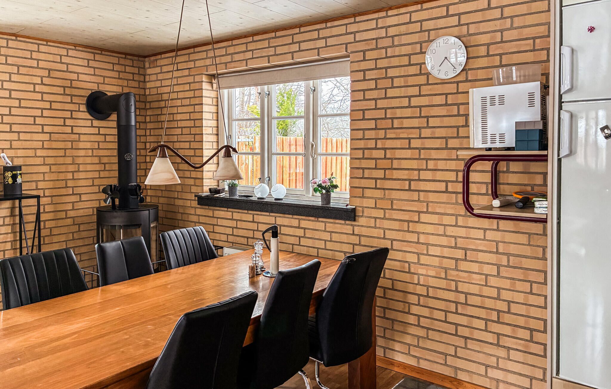 Un comedor con pared de ladrillo, mesa de madera, sillas negras, estufa negra, ventana con vistas y un reloj.
