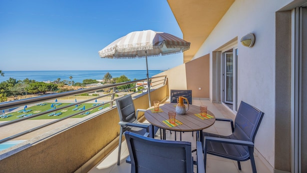 A balcony with a table, chairs, and a striped umbrella overlooking a coastal view. - Super Beach Apartment w/ Pool in the private condominium of Quinta da Barracuda (Albufeira)