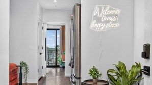 Un pasillo moderno con un letrero de neón que dice “Bienvenido a nuestro lugar feliz”, un refrigerador de acero inoxidable, una pequeña mesa redonda con una planta y vistas a un balcón con mobiliario exterior.