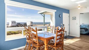 Uma área de refeições com mesa e cadeiras, uma janela grande com vista para a praia e veículos estacionados, e uma porta que dá para um quarto.
