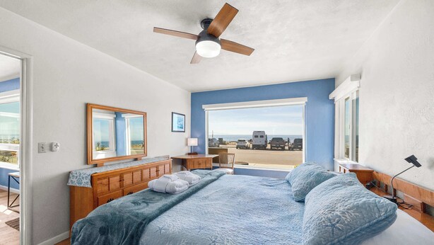 Quarto com cama, ventilador de teto, cômoda, escrivaninha, cadeira e uma grande janela com vista.