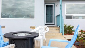 Uma área de pátio com uma mesa redonda e cadeiras, um espaço para fogueira e uma porta que dá para um alpendre coberto.