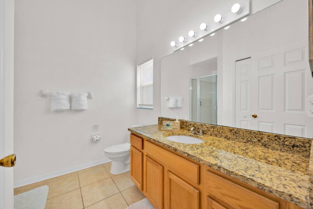 A bathroom with a white toilet, a sink with a granite countertop, wooden cabinets, and a large mirror with lights.