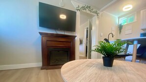 A living room with a fireplace, a flat-screen TV mounted above it, a round wooden table, and a potted plant. - Cozy Bayfield Retreat • Pool & Hot Tub • Near Grand Bend • Lake Huron Nearby 2 (Bayfield)
