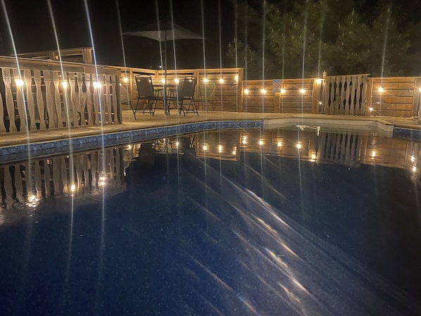 Una piscina nocturna con cubierta de madera y sillas, rodeada por una valla de madera con luces.