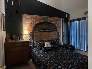 Ein Schlafzimmer mit Ziegelmuster-Kopfstütze, einem Bett mit Harry-Potter-Motiv Kissen, einer Kommode und einem Fenster mit sternenbesetzter Gardine.