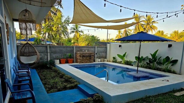Une piscine rectangulaire bordée d’une terrasse carrelée, entourée de pelouse et de plantes en pot. On y trouve des chaises bleues le long du bord de la piscine, un fauteuil suspendu et une cheminée en pierre.