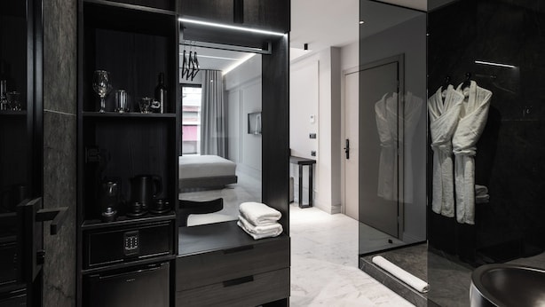 Un bagno moderno con cabina doccia in vetro, mobiletti in legno scuro e un letto visibile sullo sfondo.