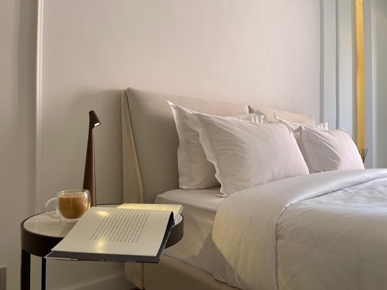 A neatly made bed with white linens, a lamp, a cup of coffee, and a book on a bedside table.