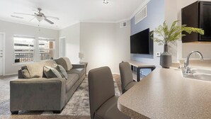 Una sala de estar moderna con un sofá, sillas, un televisor y una zona de cocina con un lavamanos y un florero.