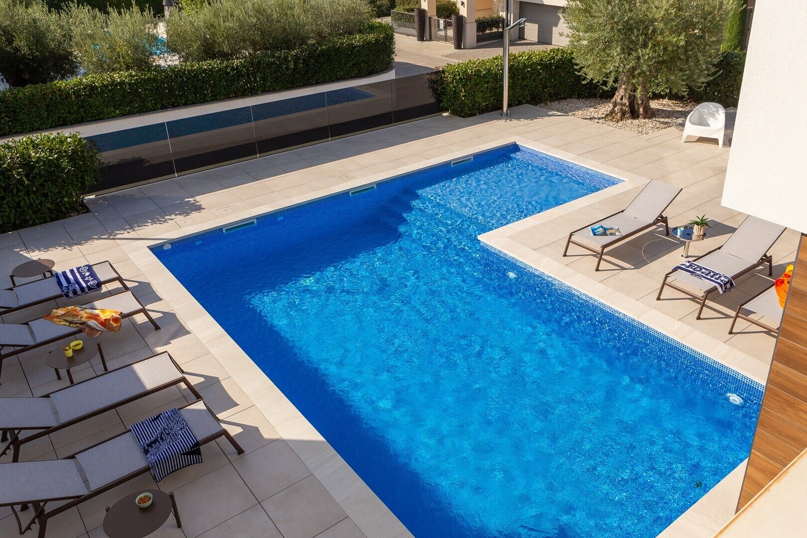 A rectangular swimming pool with clear blue water, surrounded by lounge chairs and sunbeds, and a small table with a bowl.