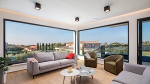Ein geräumiges Wohnzimmer mit großen Fenstern, einem grauen Sofa, einem kleinen runden Tisch und zwei Sesseln.