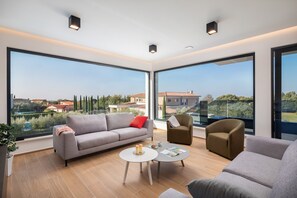 Ein geräumiges Wohnzimmer mit großen Fenstern, einem grauen Sofa, einem kleinen runden Tisch und zwei Sesseln.