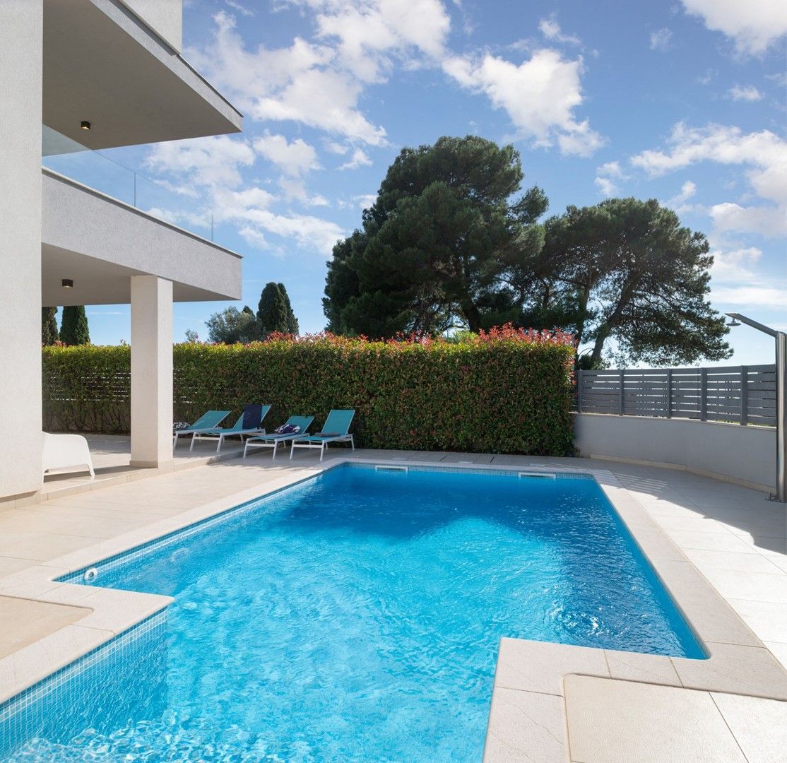 Piscina retangular com água azul cristalina, ladeada por um deck de azulejos brancos. Há espreguiçadeiras e um sebe ao fundo.
