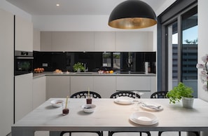 Une cuisine moderne avec un coin repas, comprenant un grand îlot central, des luminaires suspendus noirs et une vue sur l’extérieur.