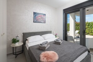 A modern bedroom with a grey bed, white pillows, a grey blanket, a fluffy pink pillow, a painting on the wall, and a view of a balcony with outdoor furniture. - Villa Fasana 3 - luxury apartment with sea view and private pool for 6 persons. (Fažana)