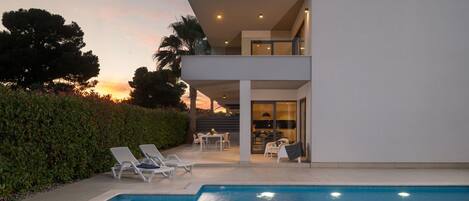 A modern two-story house with a swimming pool, outdoor seating, and a view of the sunset.