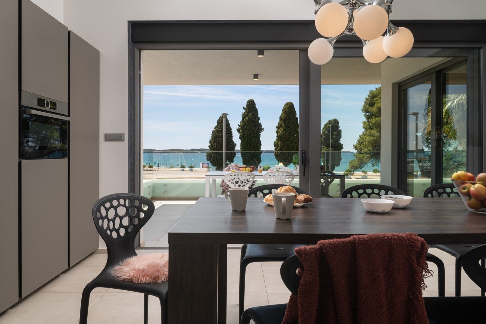 A modern kitchen with a large dining table, chairs, and a view of the sea.