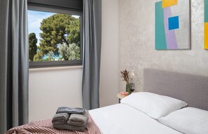 Une chambre d’hôtel avec un lit, une fenêtre avec des rideaux, une table de chevet avec un vase et une bougie, et un mur orné d’un tableau aux motifs géométriques.