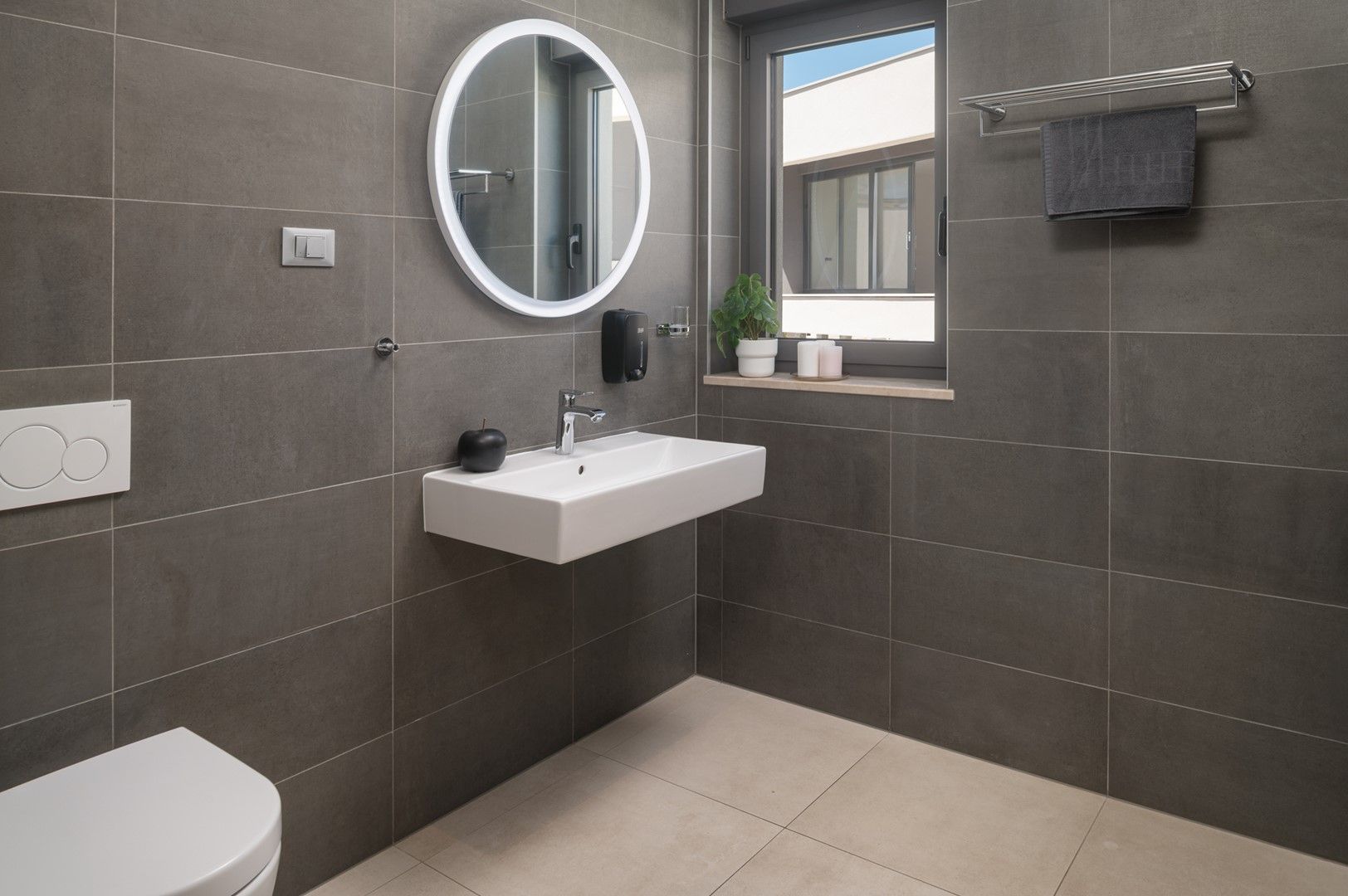 Une salle de bain moderne avec un lavabo blanc, un miroir rond, un porte-serviettes et une fenêtre.