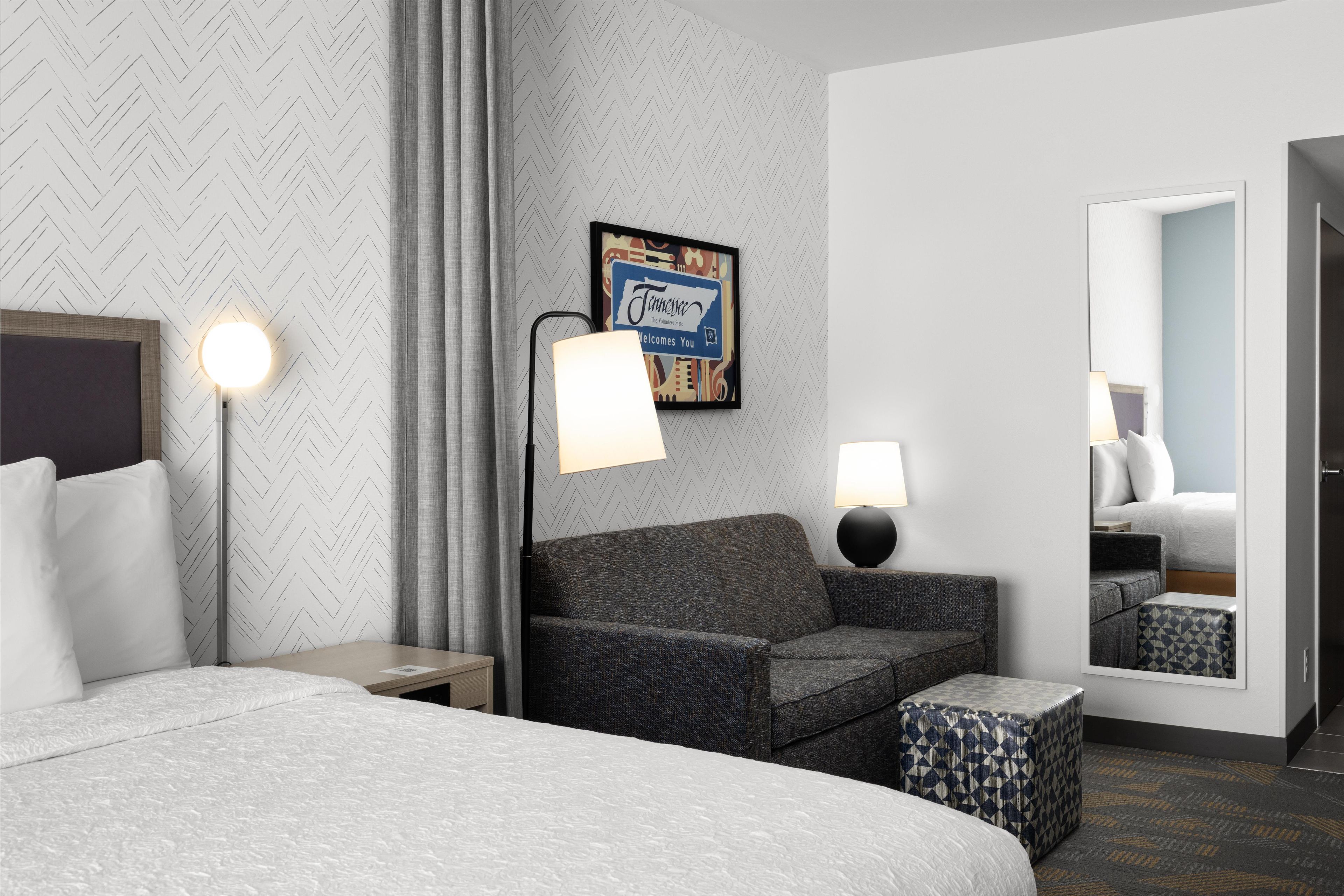 Une chambre d’hôtel comprenant un lit, un canapé, une petite table, une lampe, un miroir et un mur orné d’un motif en chevrons.