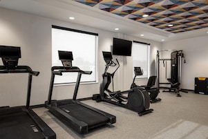 Un gimnasio con cintas de correr, elípticas, un banco de pesas y un televisor en la pared.