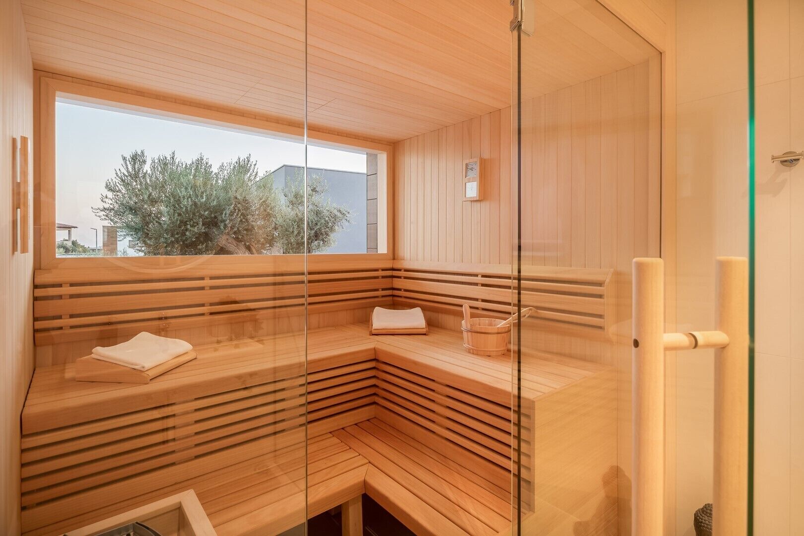 A sauna with wooden benches, a glass partition, and a window providing a view of trees and buildings.