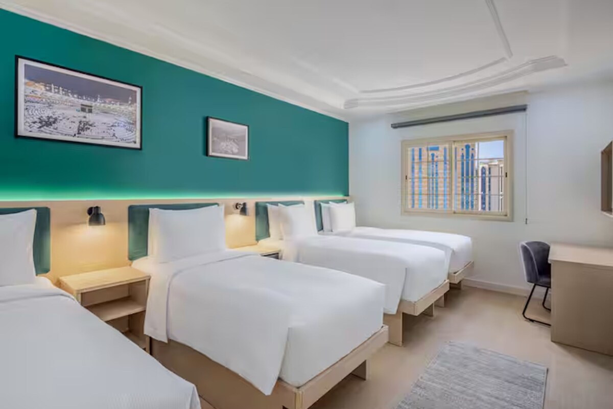 Habitación de hotel con tres camas, un escritorio y un ventanal con vistas a edificios.