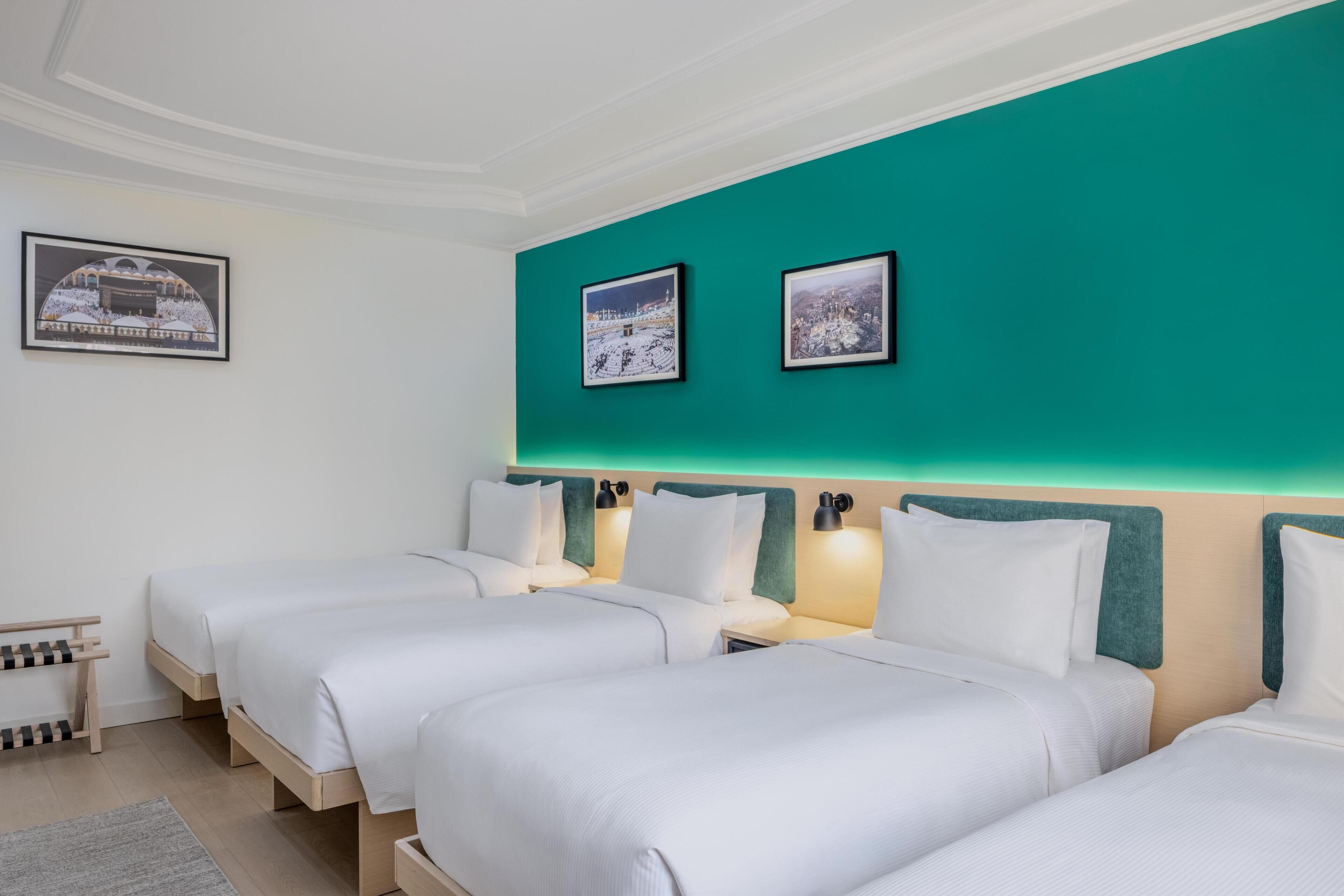 Habitación de hotel con tres camas, paredes de acento color aguamarina, cuadros enmarcados y una pequeña zona de estar.