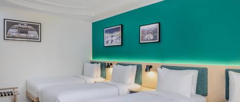 Habitación de hotel con tres camas, paredes de acento color aguamarina, cuadros enmarcados y una pequeña zona de estar.