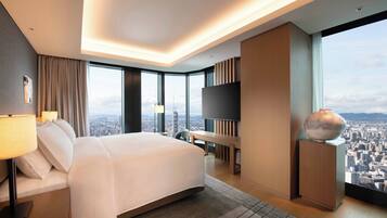 A modern hotel room with a large bed, a flat-screen TV, a city view, and a large vase on a pedestal.