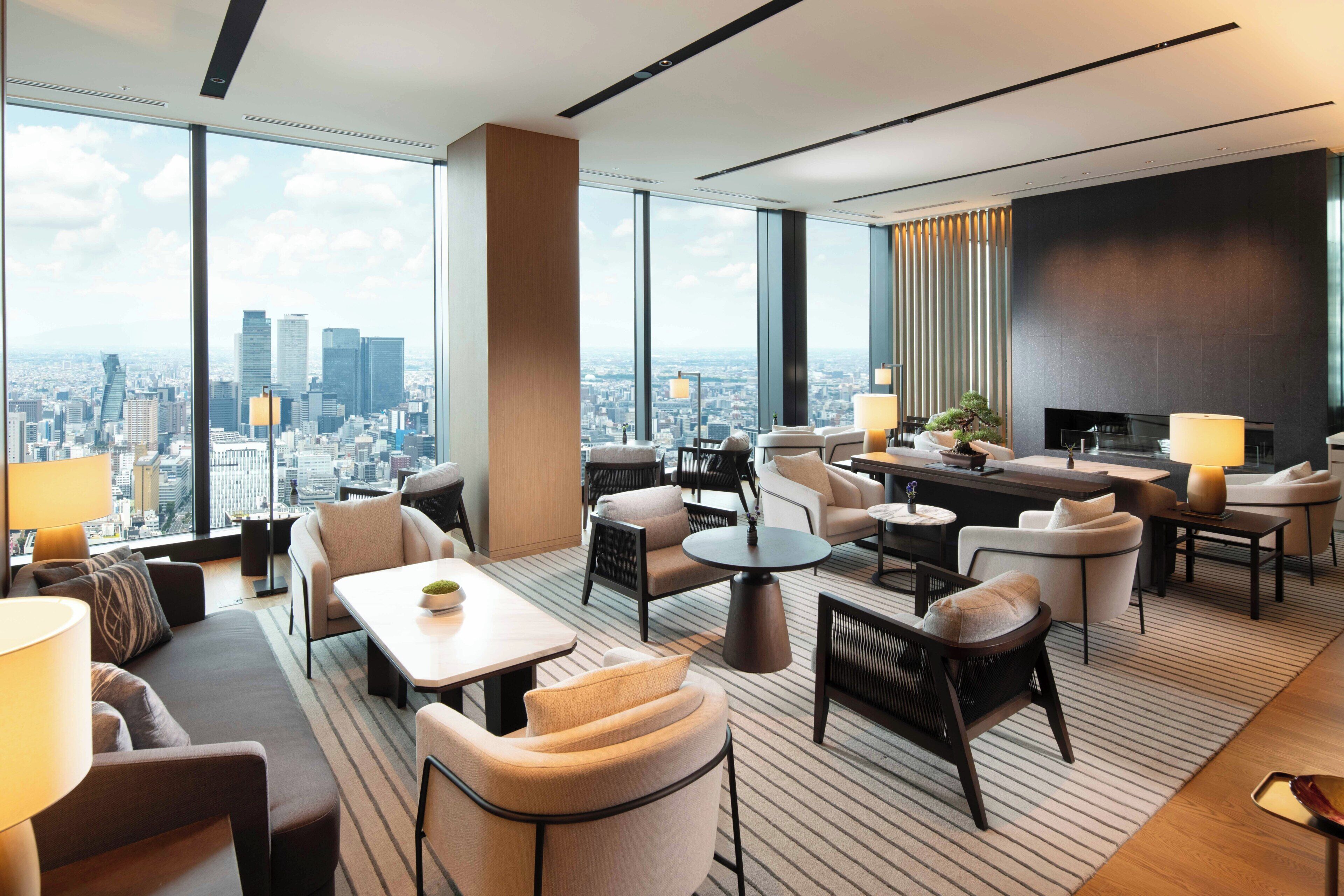 A modern hotel room with a city view, a seating area with sofas and chairs, a dining table, and a TV.