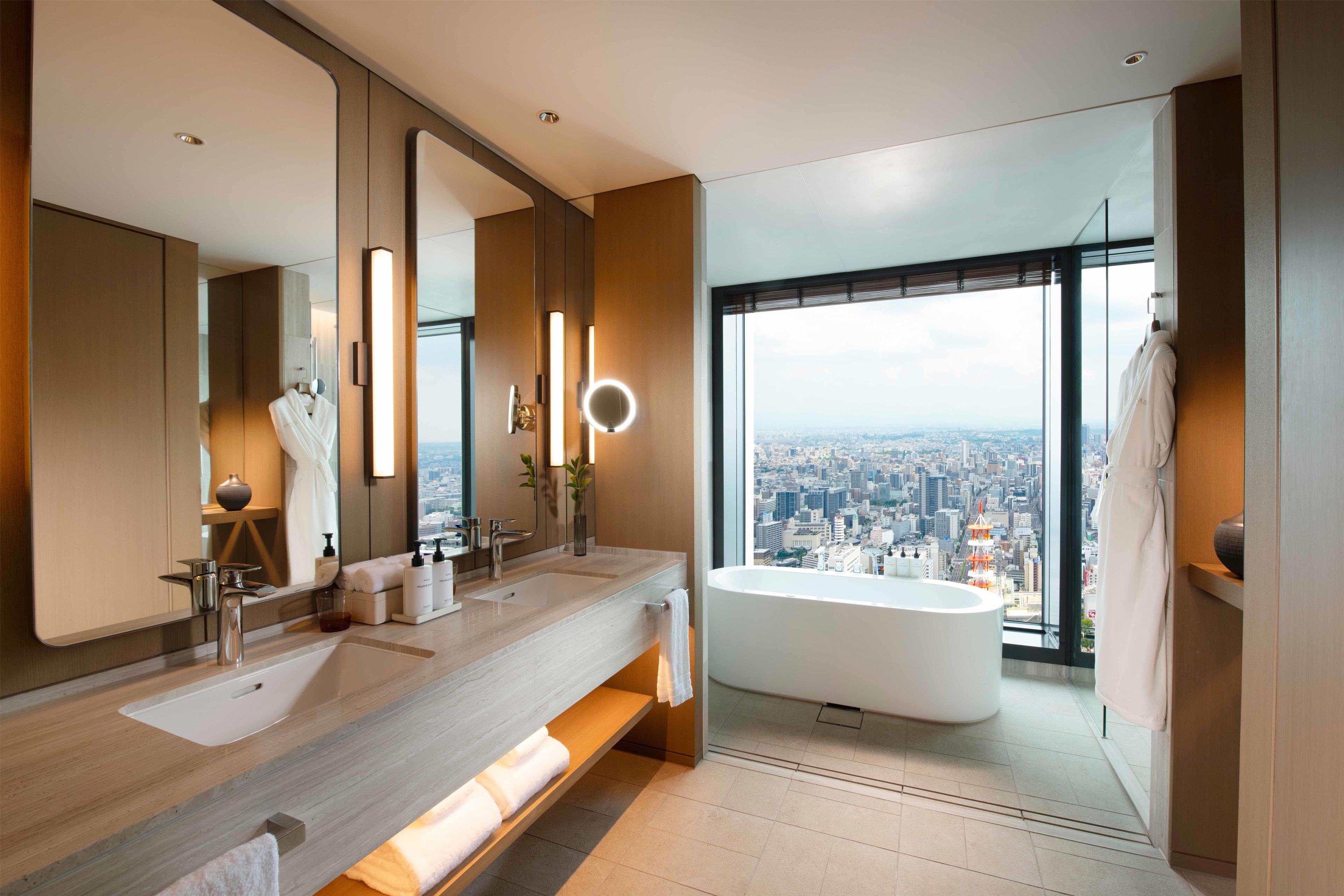 Une salle de bain moderne avec vue sur la ville, un grand miroir, deux lavabos, une baignoire et un meuble-lavabo avec des articles de toilette.