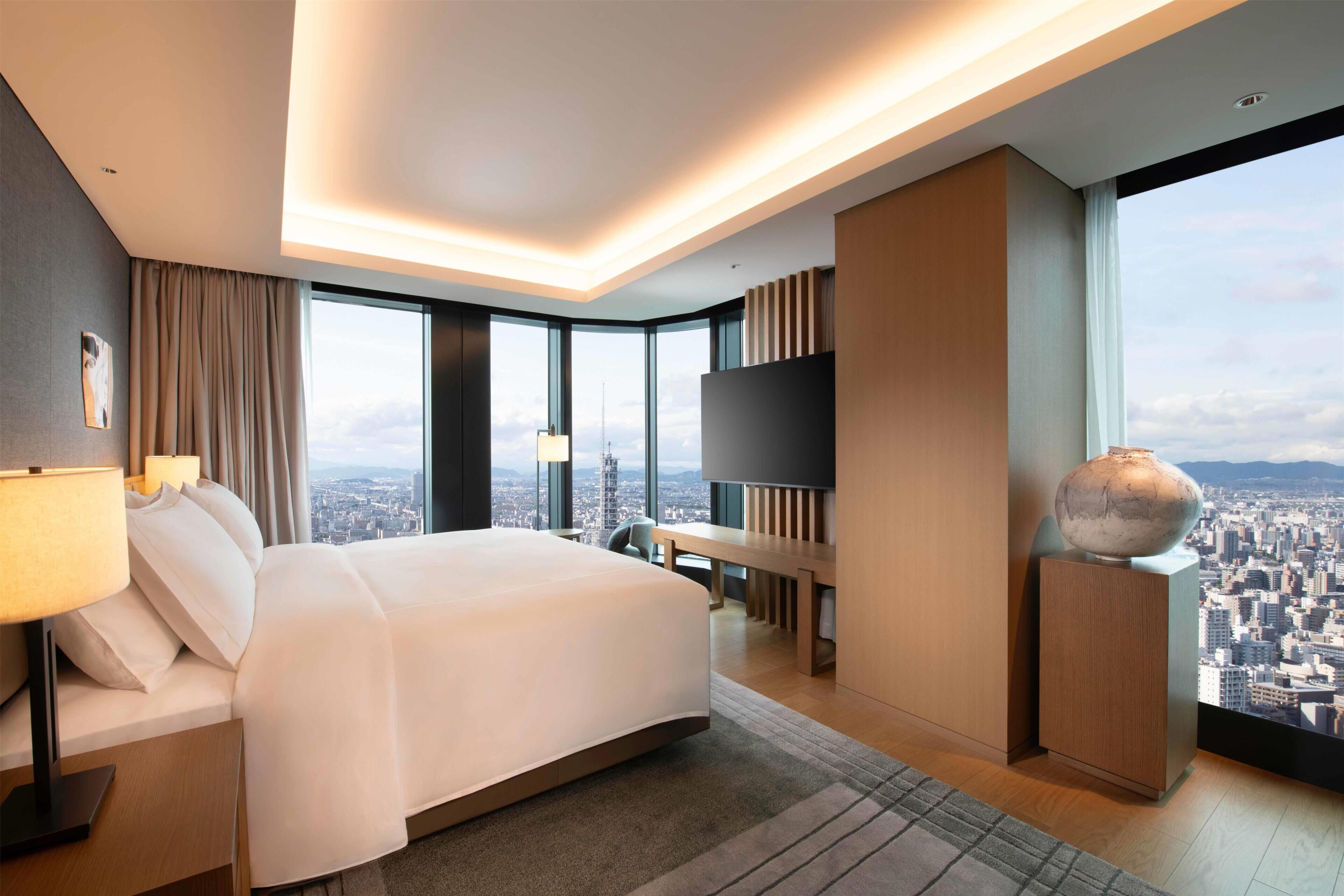 A modern hotel room with a large bed, a flat-screen TV, a city view, and a large vase on a side table.