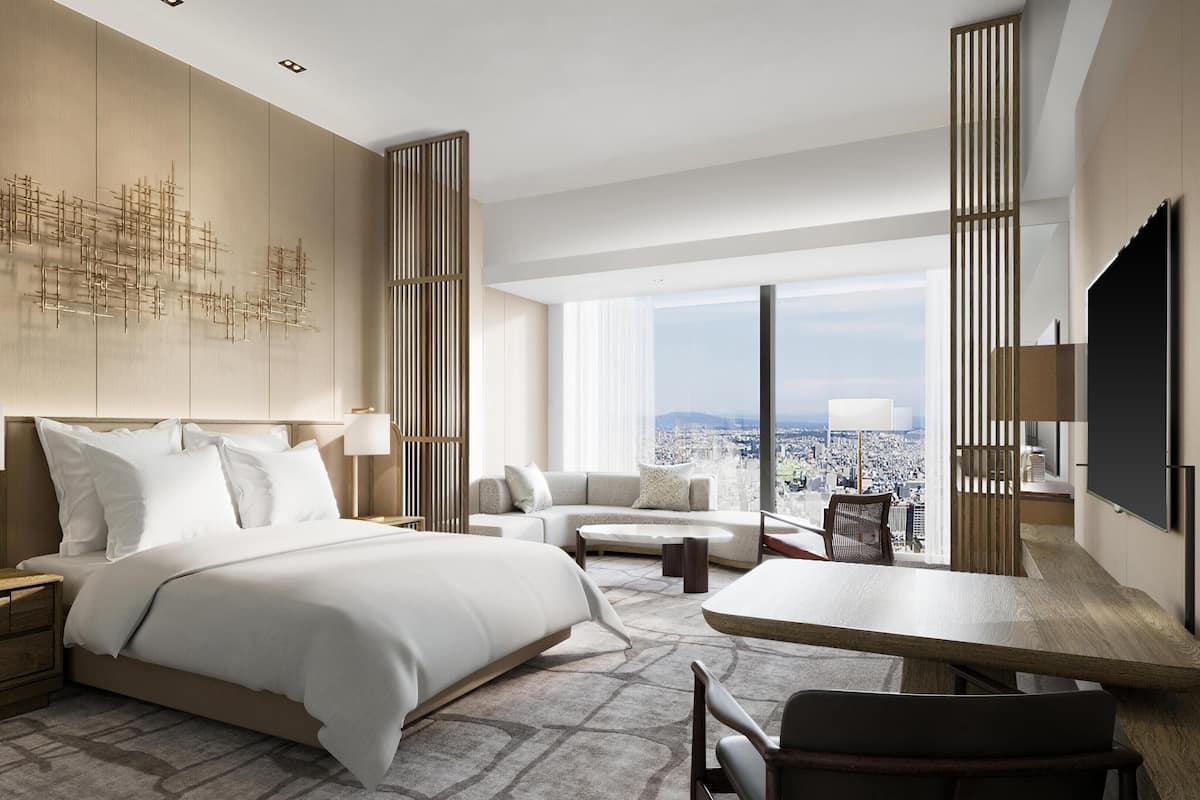 A modern hotel room with a large bed, a sofa, a dining table, and a city view.