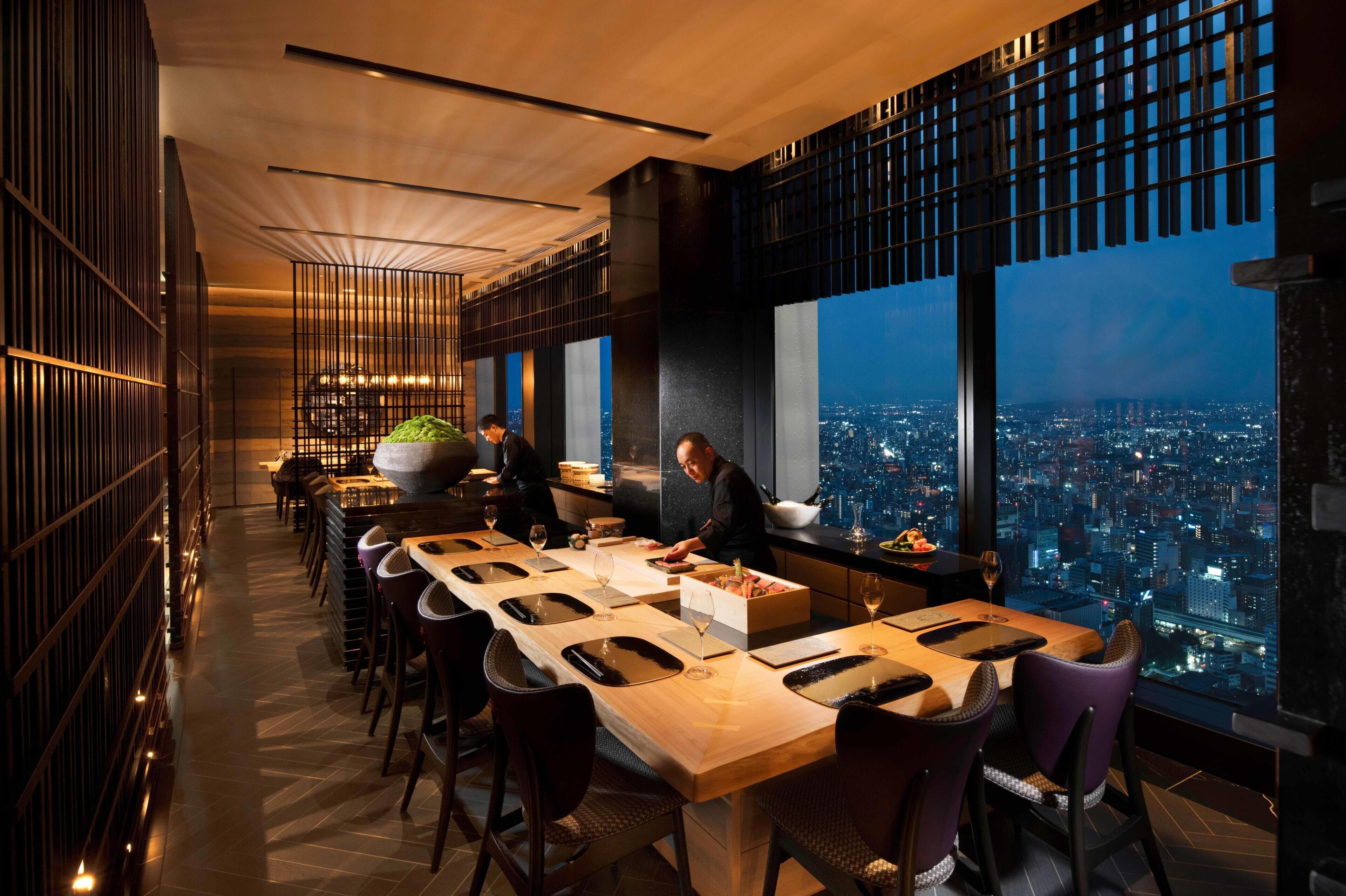 Un espace de restauration moderne offrant une vue sur la ville, avec une longue table et des chaises, un chef cuisinier en train de préparer les plats et une grande fenêtre donnant sur le paysage urbain.