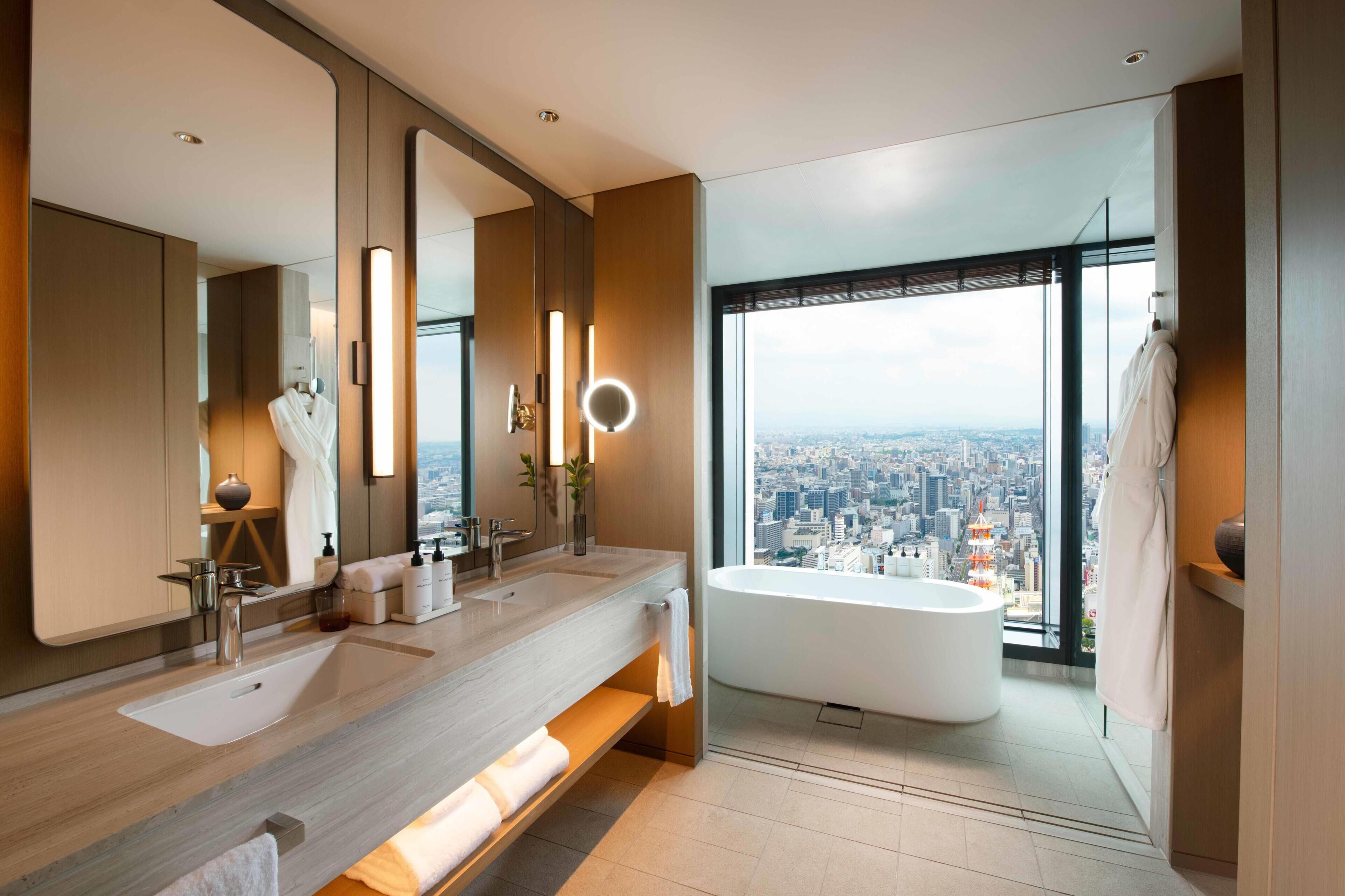 Un baño moderno con vistas a la ciudad, un espejo grande, dos lavamanos, una bañera y un tocador con artículos de aseo.