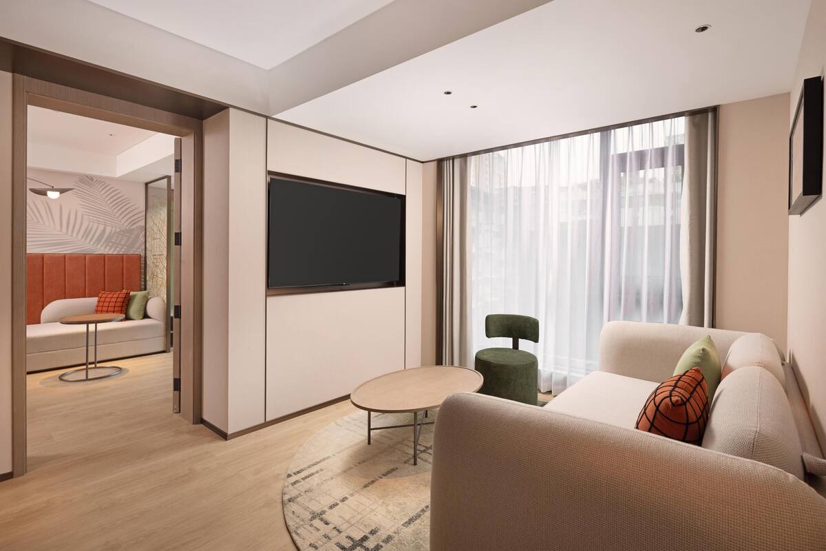 Una habitación de hotel moderna con sofá, una mesita, televisor y un ventanal grande.