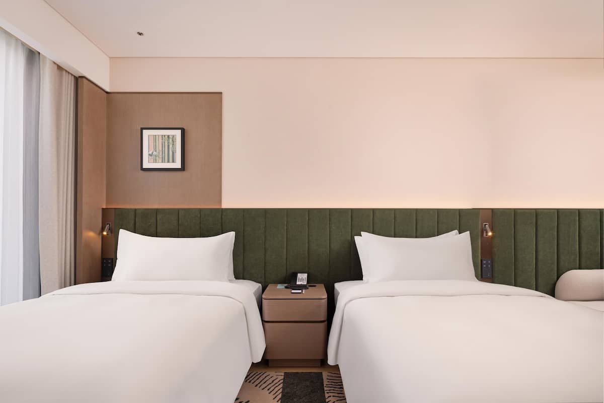 Habitación de hotel con dos camas, una mesita de noche con teléfono, un cuadro en la pared y una ventana con cortinas.