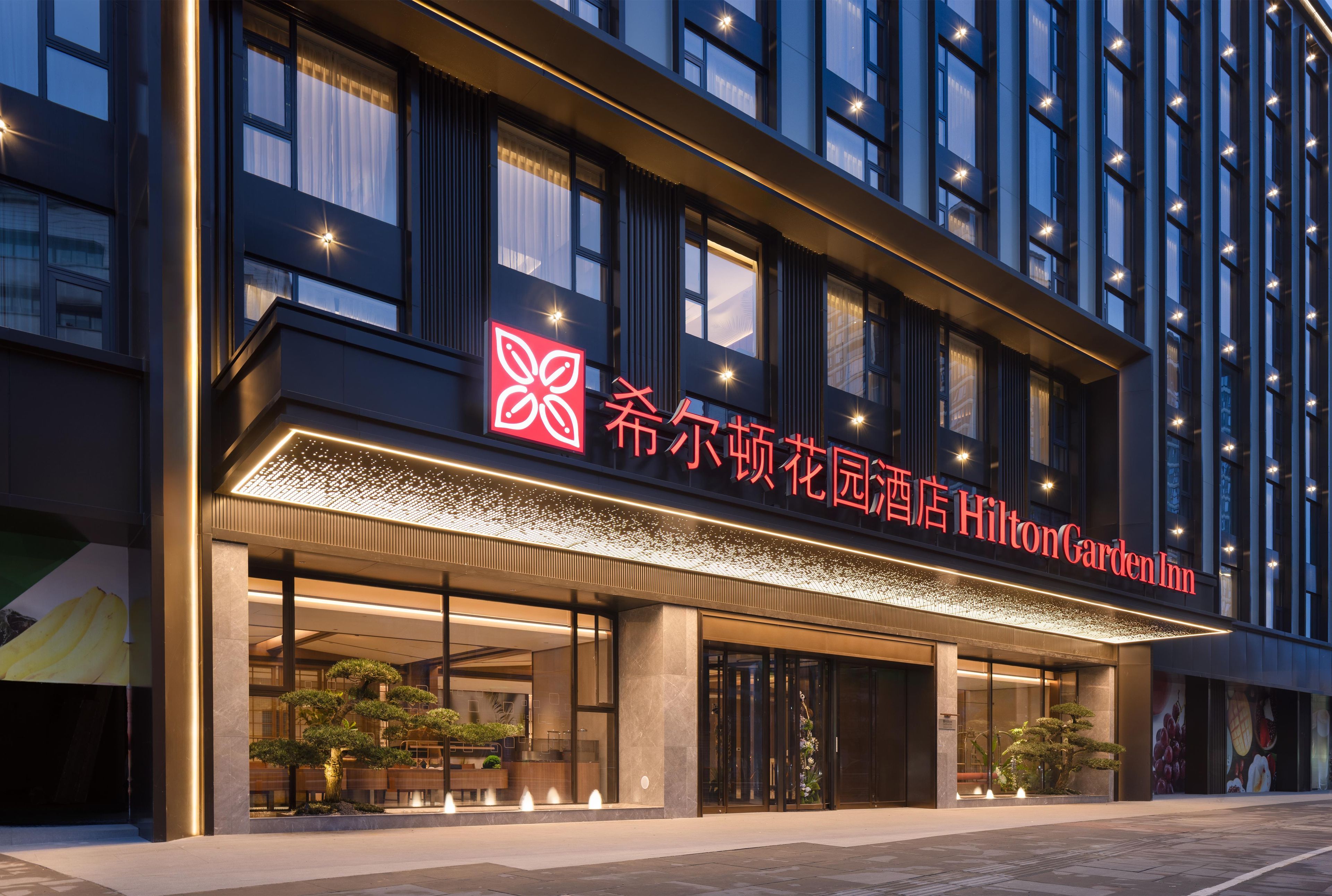 L’extérieur d’un hôtel au design moderne, avec une enseigne bien visible affichant des textes en chinois et en anglais, de grandes fenêtres et une entrée bien éclairée.