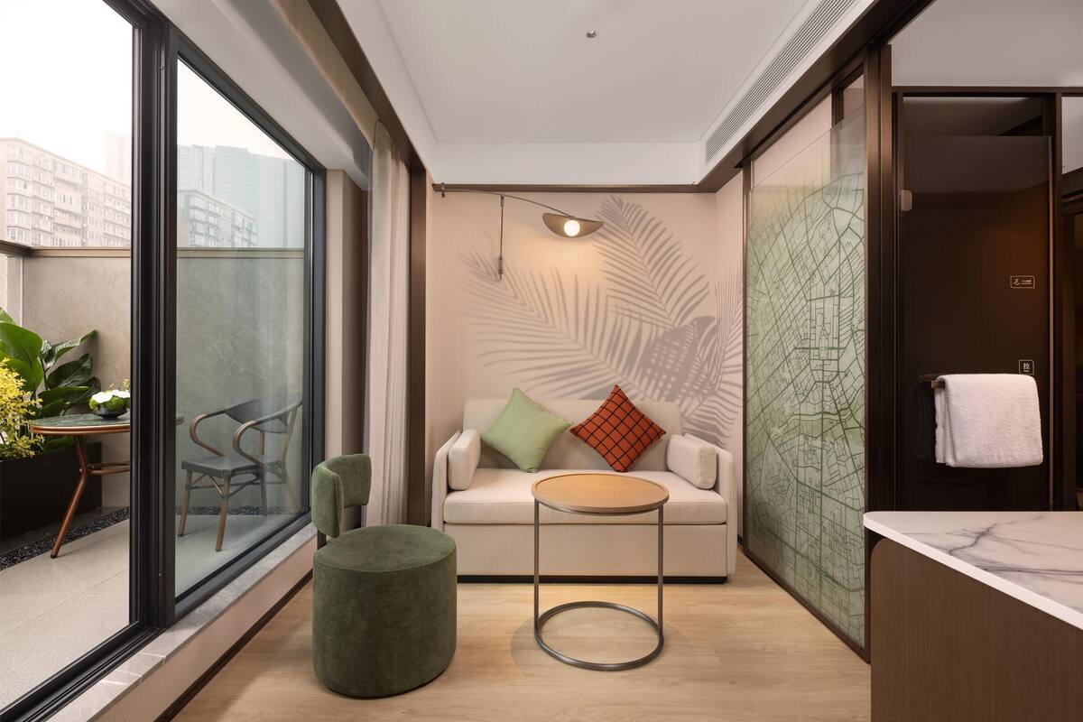 Una habitación de hotel moderna con un ventanal grande, un sofá con cojines, una mesa redonda, un otoman verde y un baño con mampara de vidrio.