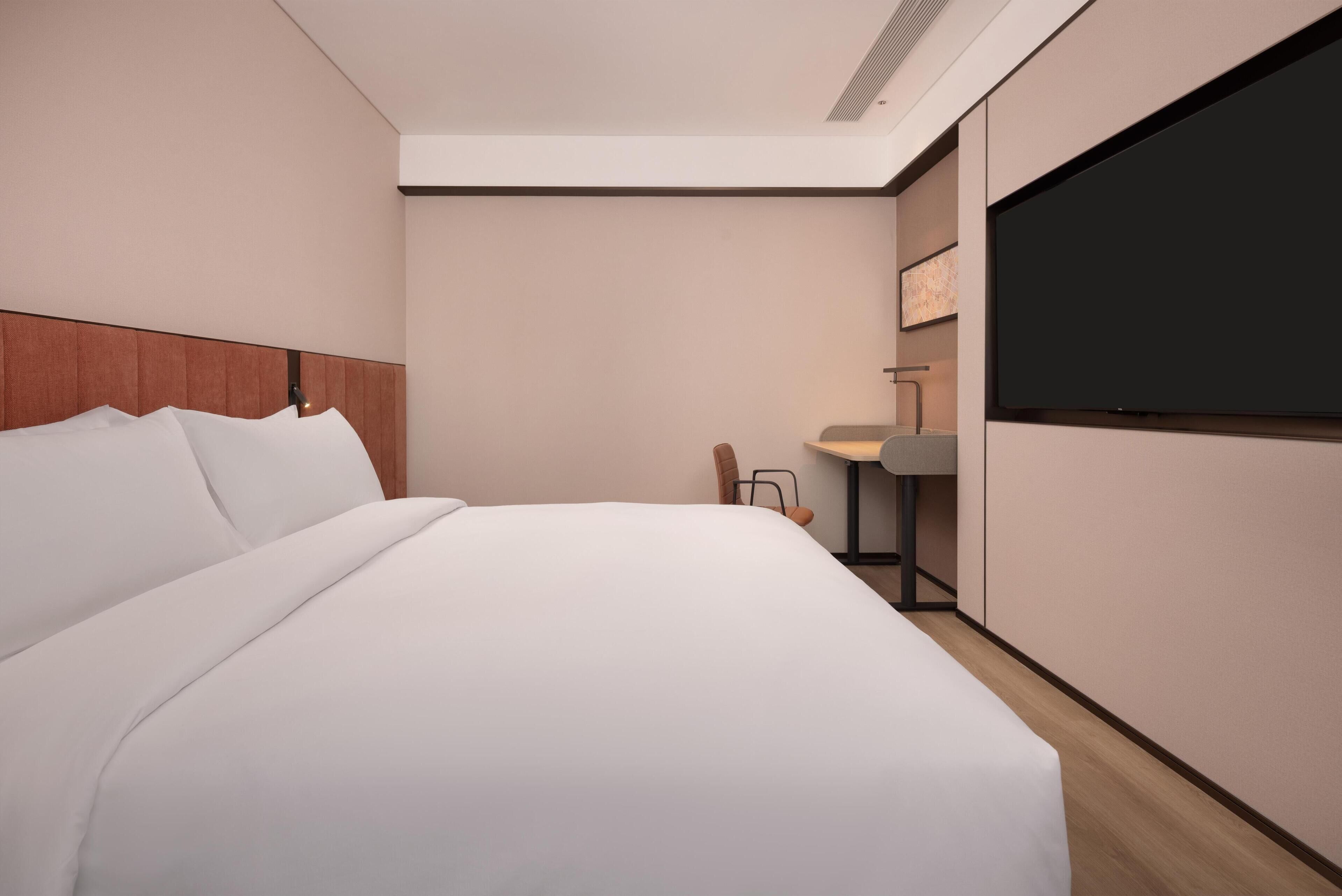 A modern hotel room with a large bed, a flat-screen TV, a desk with a chair, and a wall-mounted TV.