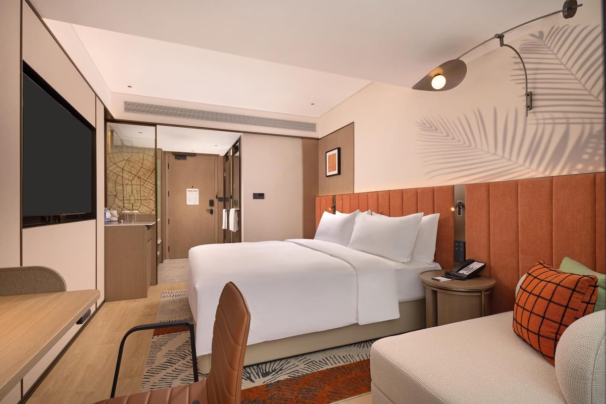 Habitación de hotel con cama, escritorio, silla, televisión, mesita de noche con teléfono y lámpara de pared.