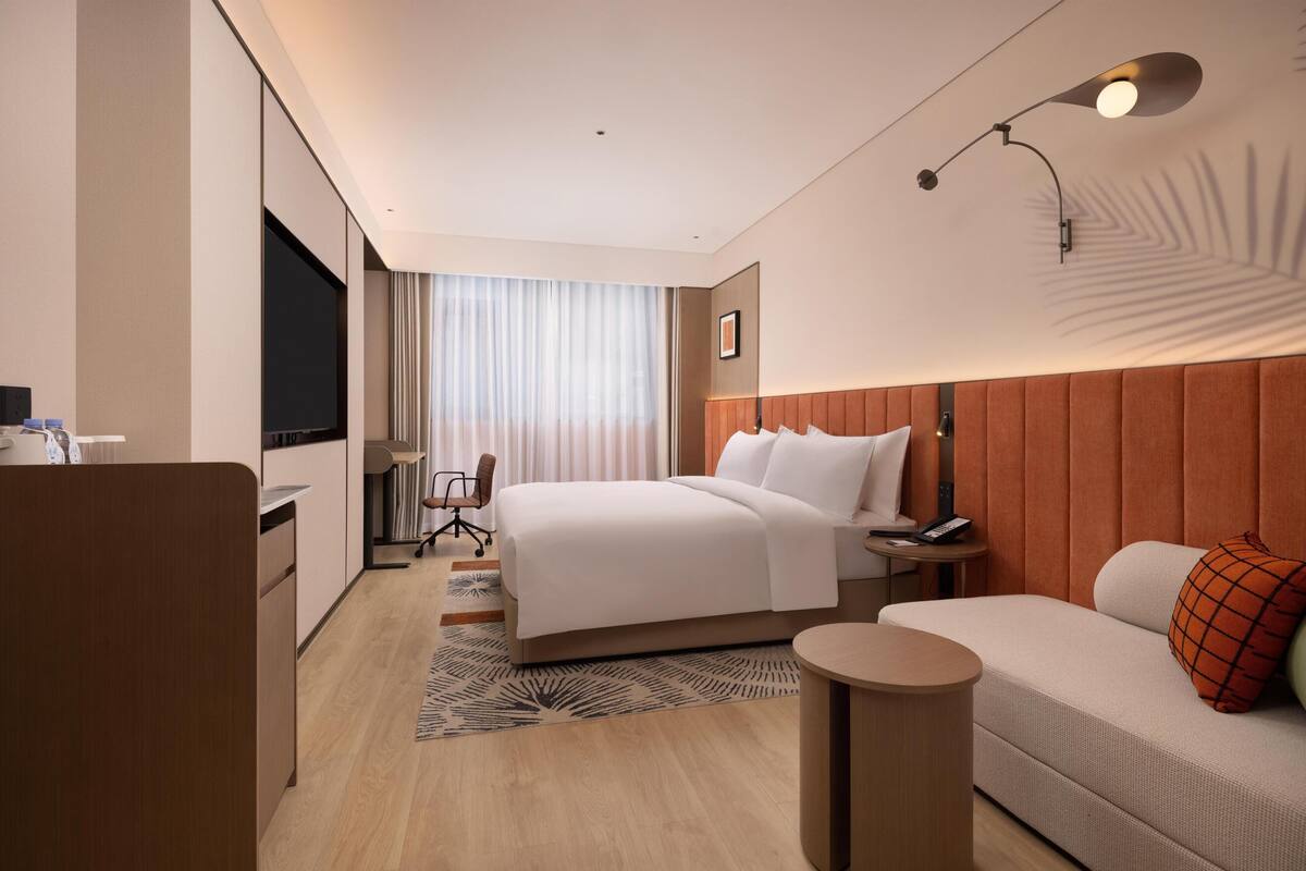 Habitación de hotel con cama, sofá, escritorio, silla, televisión y ventana con cortinas.